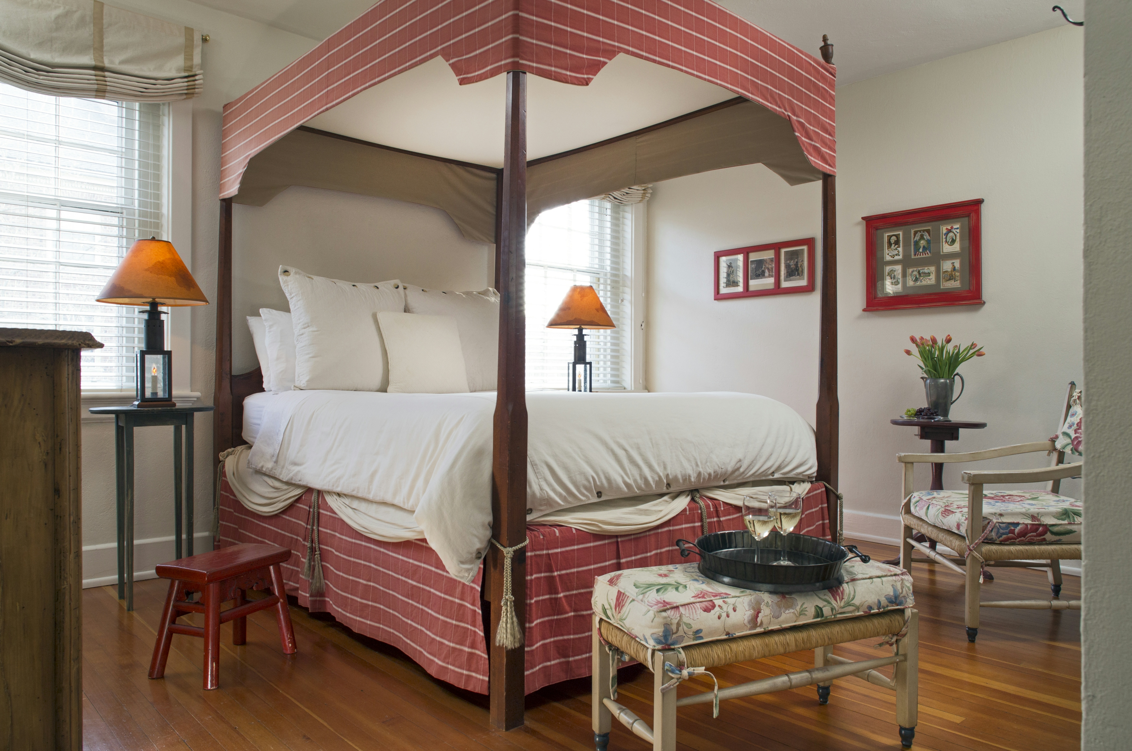 Lofty four-poster canopy bed with nightstands and lamps, hardwood floor, bench and chair with matching floral cushions