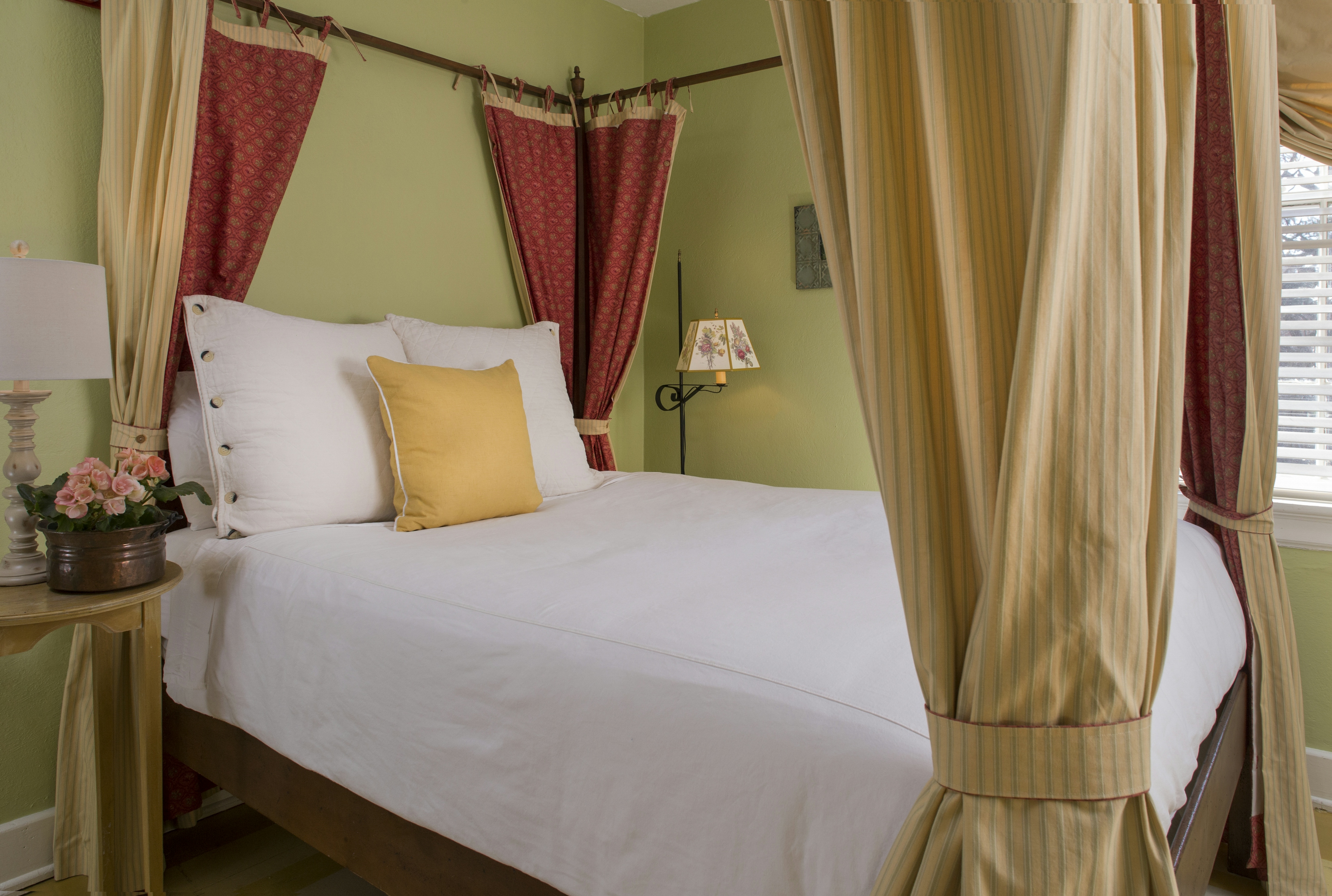 Four-poster canopy bed with red and gold drapes, nightstand with table lamp and flowers