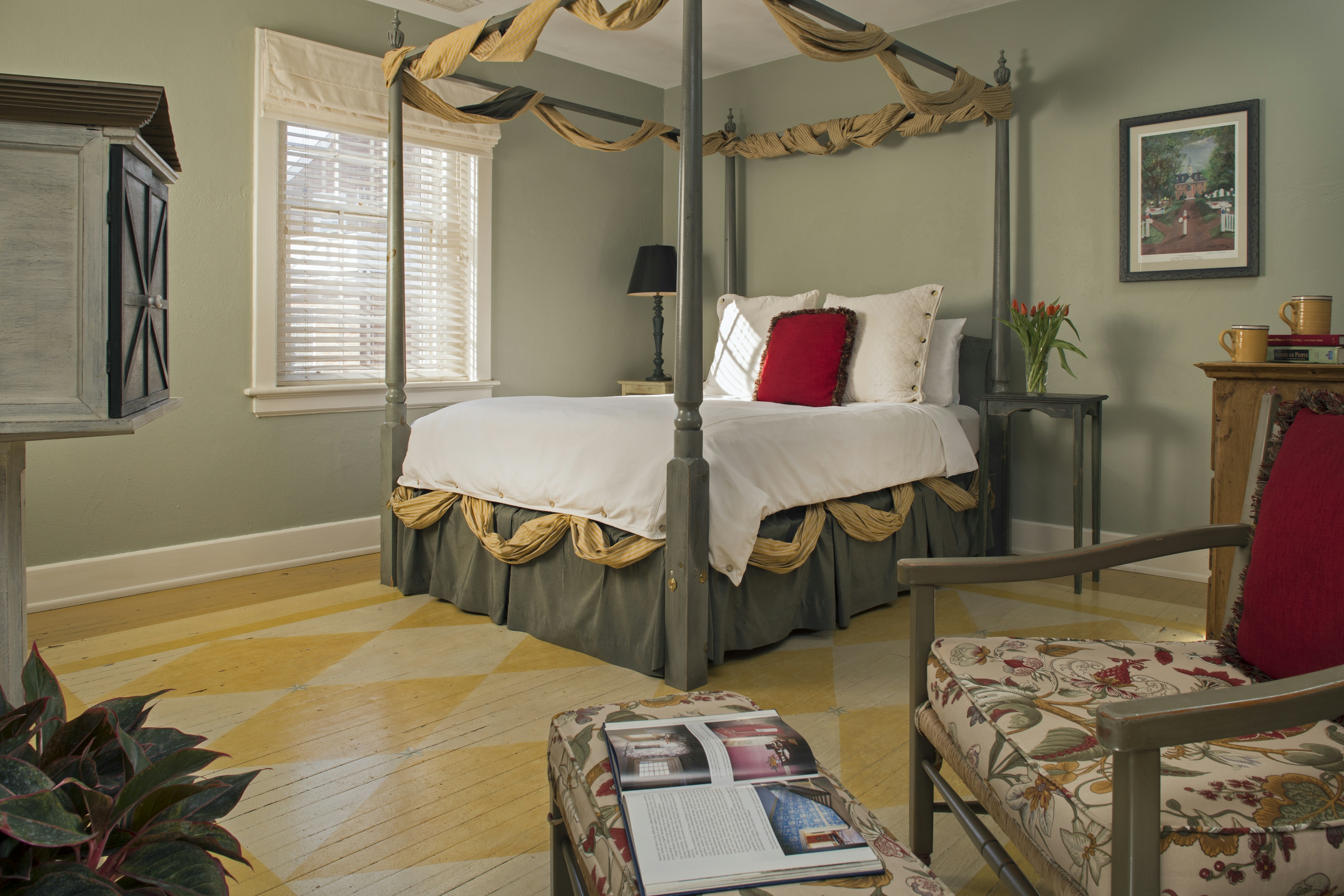 Painted wood four-poster bed swagged at the top with gold fabric, wooden chair with floral cushions and matching footstool in foreground