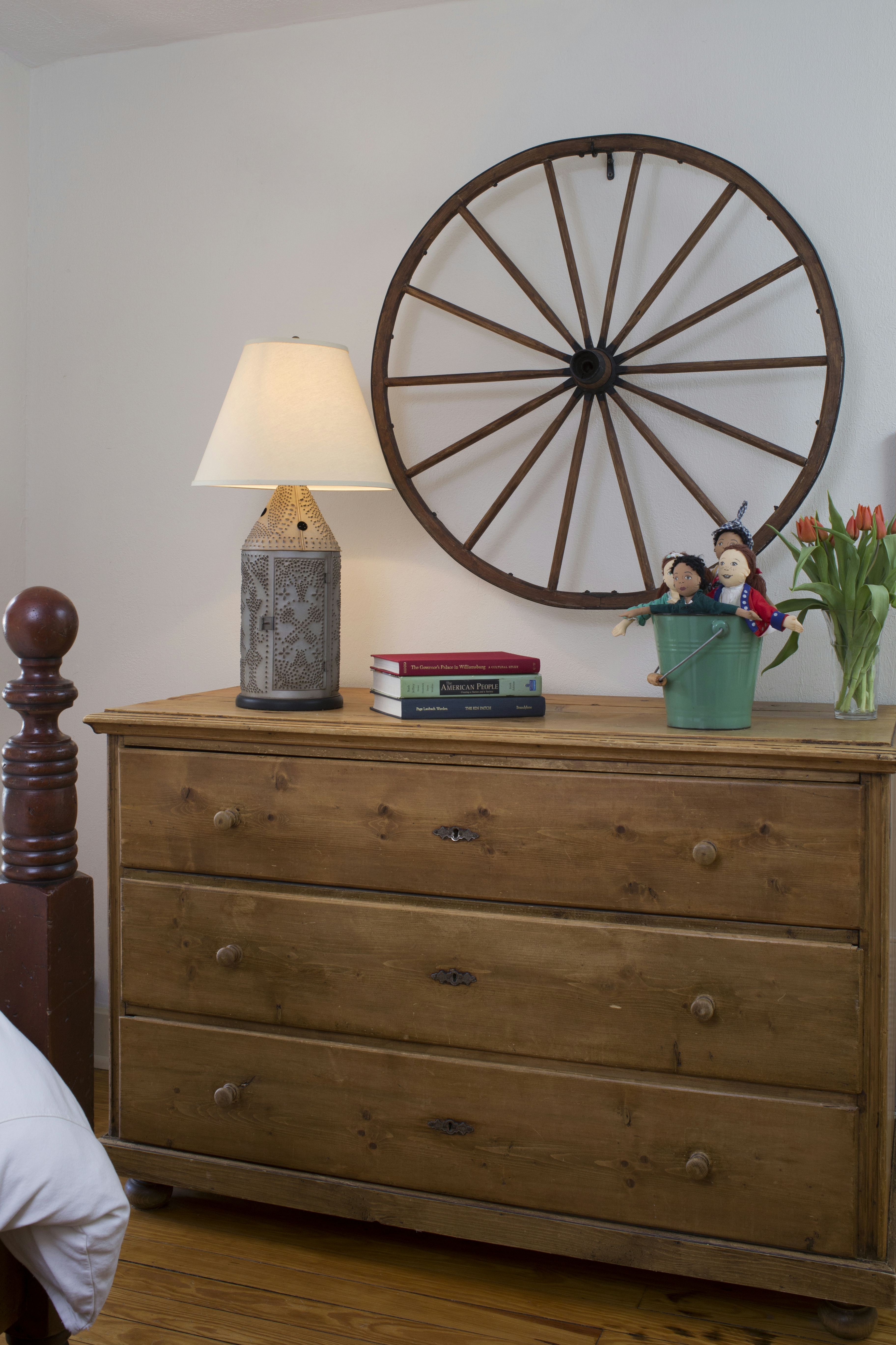 Wooden chest of drawers with table lamp, books, vase of tulips, wagon wheel hanging on wall above