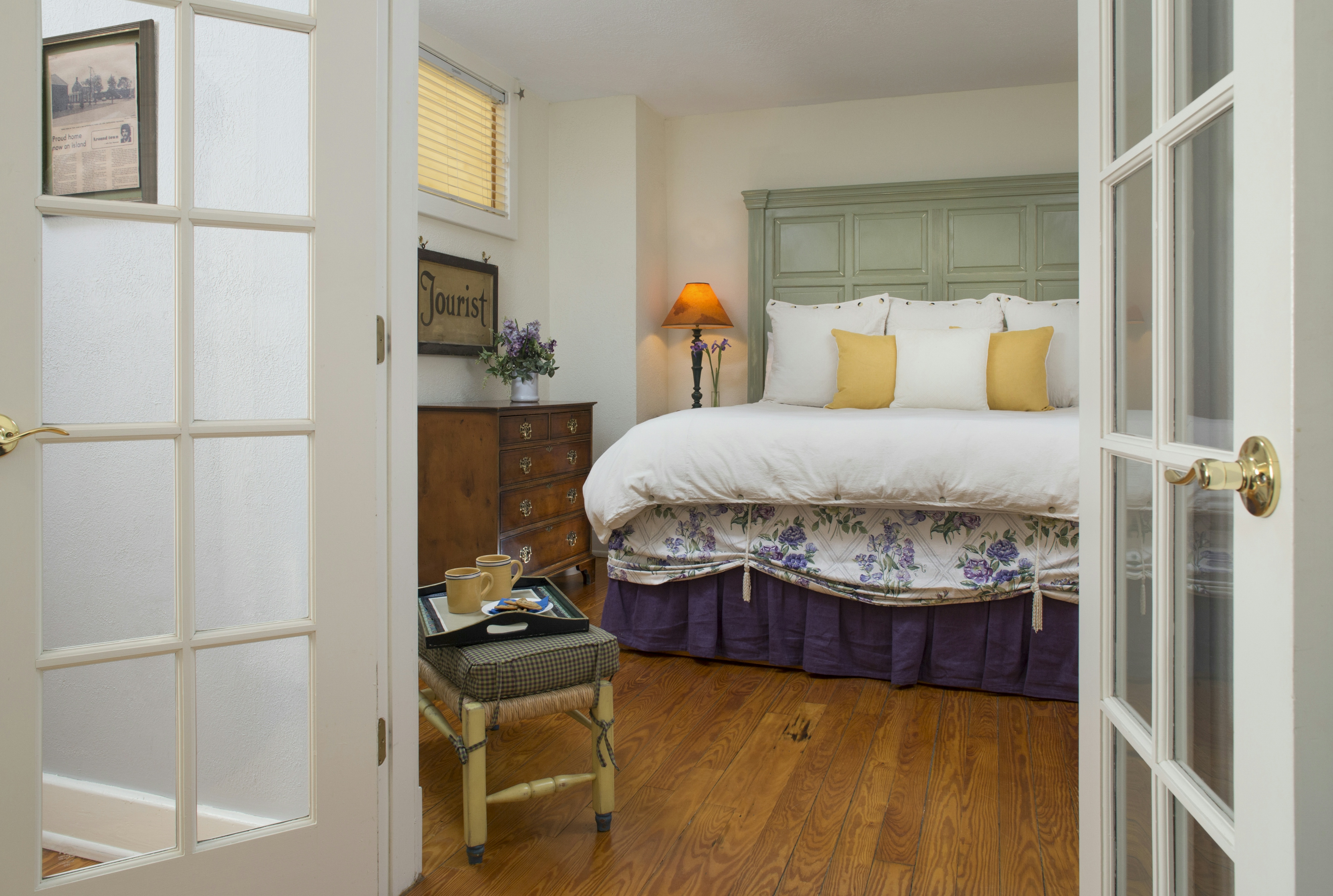 View from doorway into bedroom, beautifully appointed bed with high painted headboard, nightstand with lamp, dresser