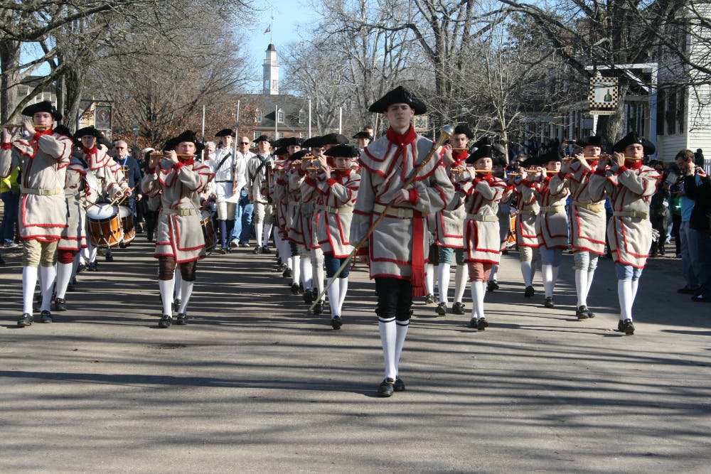 A fife and drum corp dressed in 18th century attire walking down a street playing their instruments.