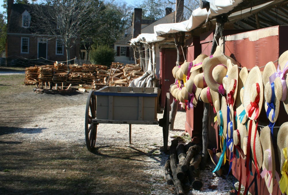 Colonial Brick Building in background, piles of chopped wood, old wagon and hanging ladies straw hats with multi color ribbons