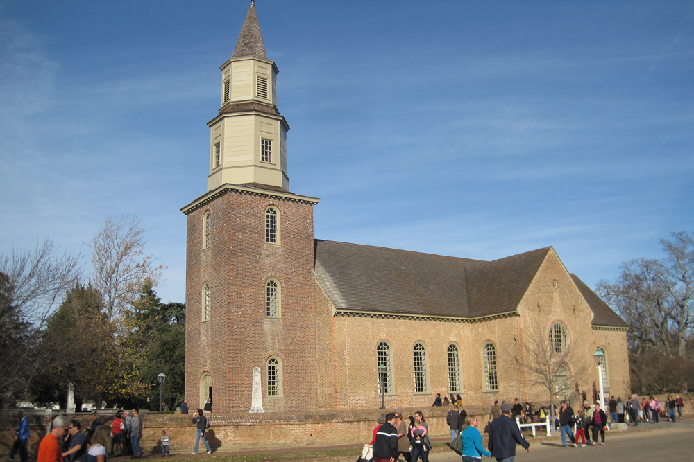 Tall yellow brick church with white steeple with many people walking about.