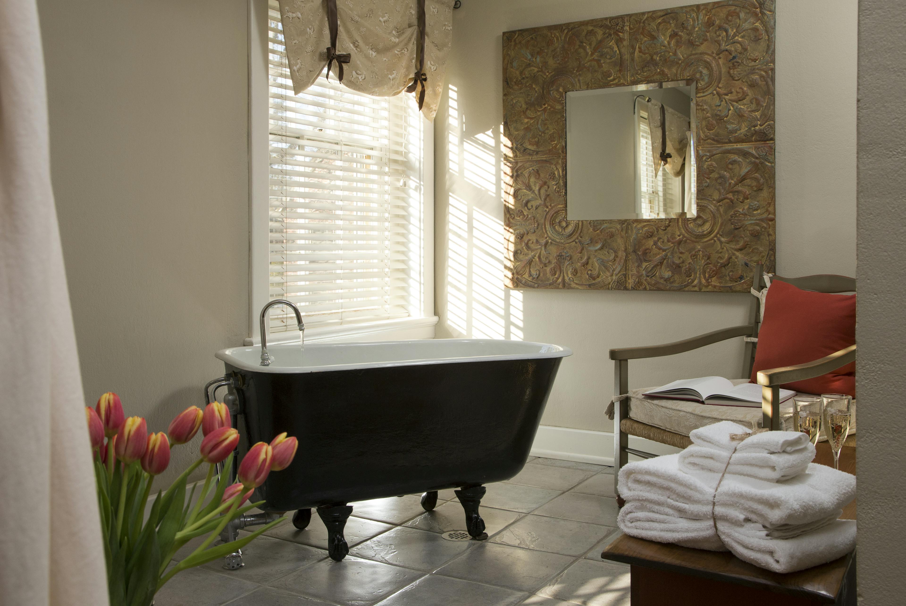 Clawfoot soaking tub on tiled floor, sunny window with blinds and valance, ornately framed mirror, and comfortable chair