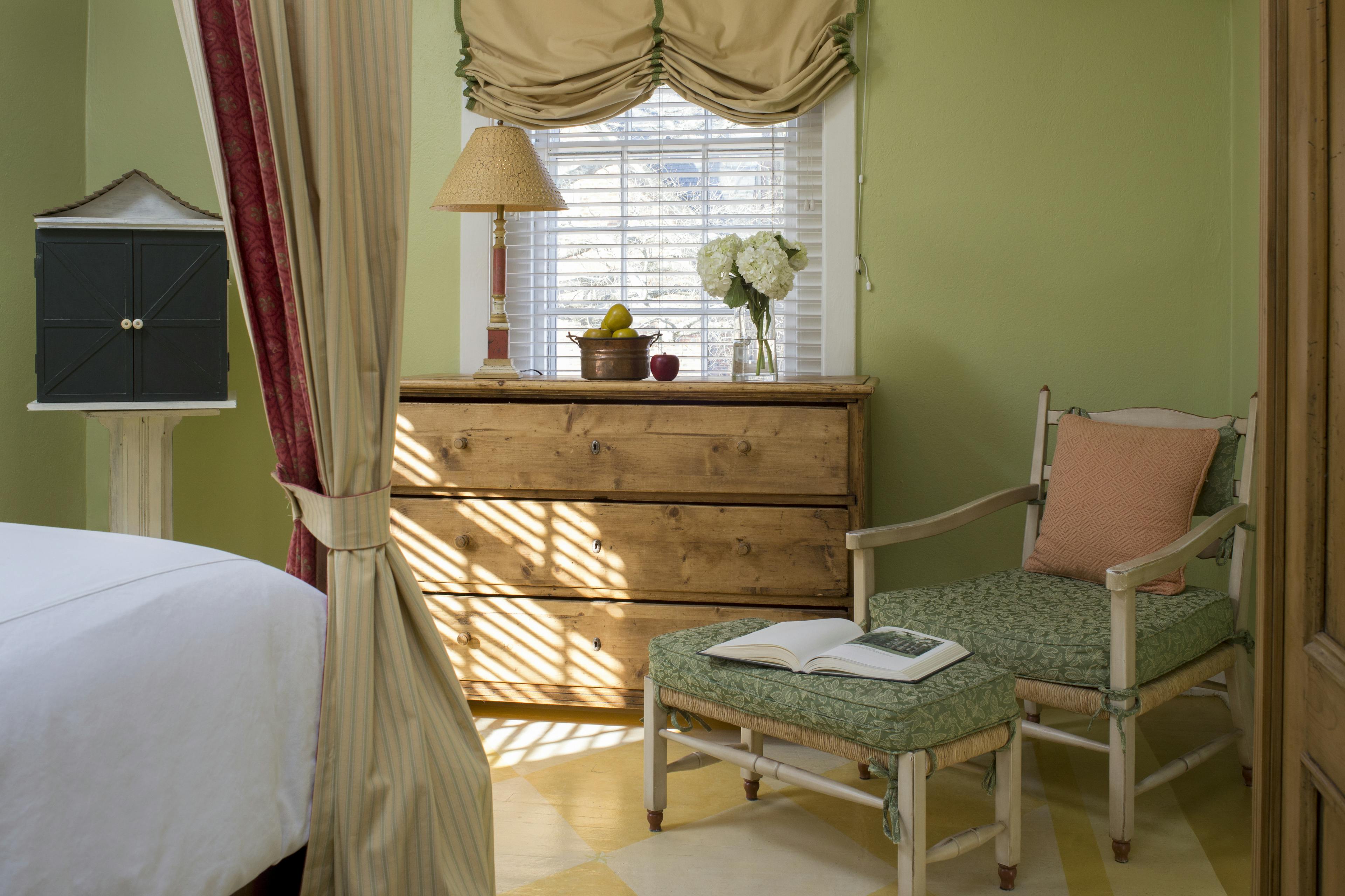 Wooden chair with cushions and matching footstool at foot of bed, wooden chest of drawers with lamp and vase of flowers