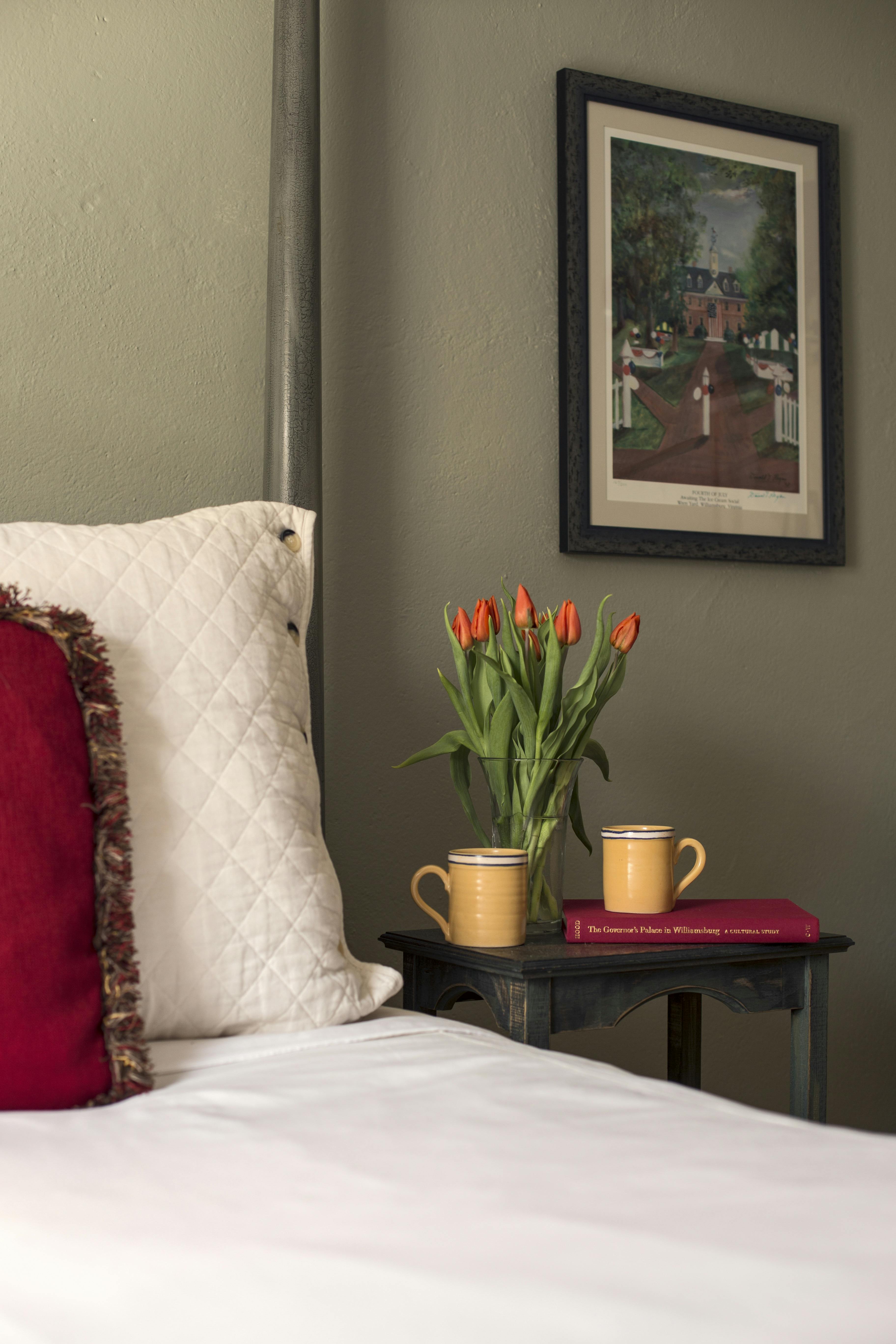 Small bedside table with two yellow mugs, a book, and vase with tulips, painting of William & Mary campus on wall