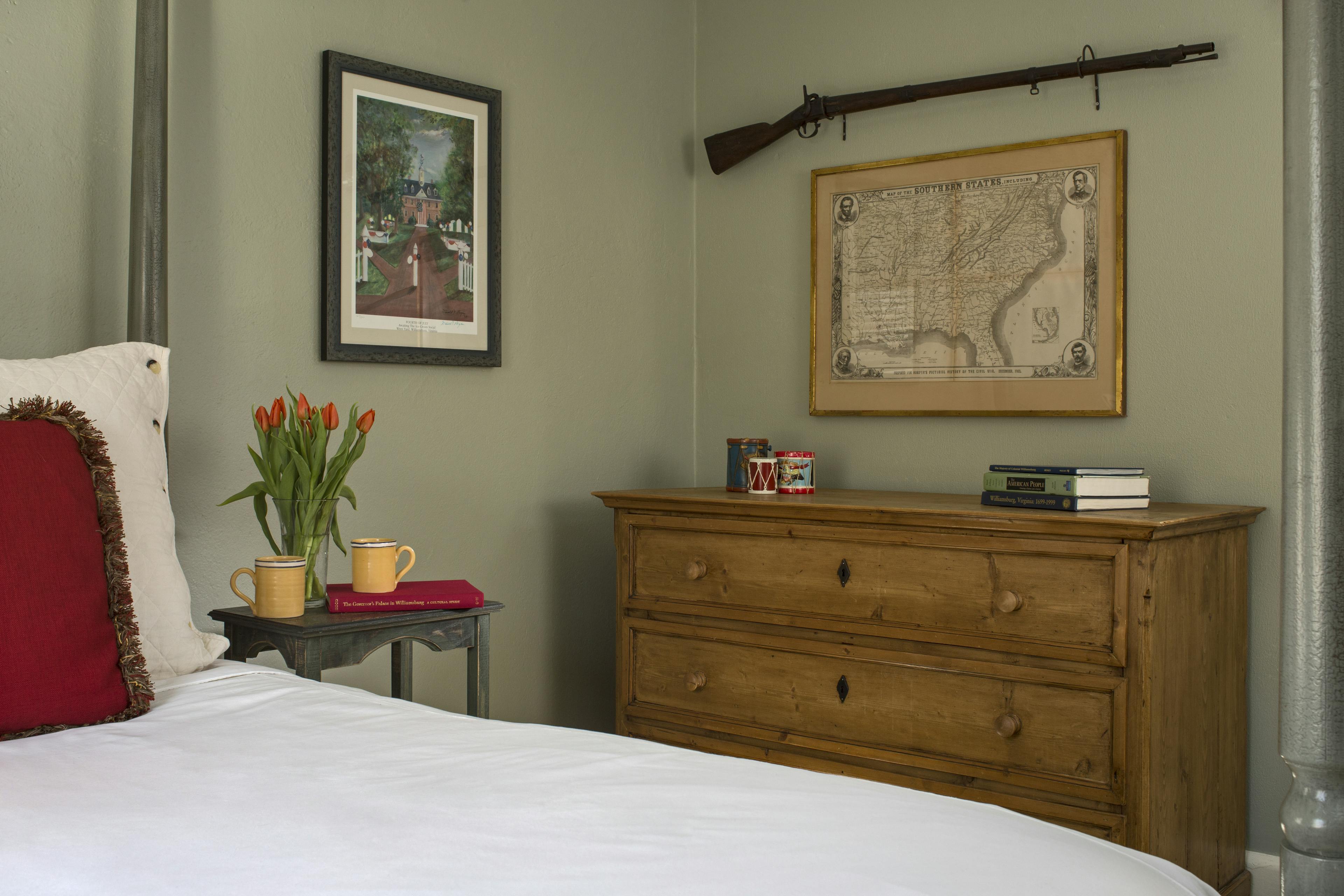 Partial view of four-poster bed, bedside table, large bureau with map and long rifle hanging on wall above