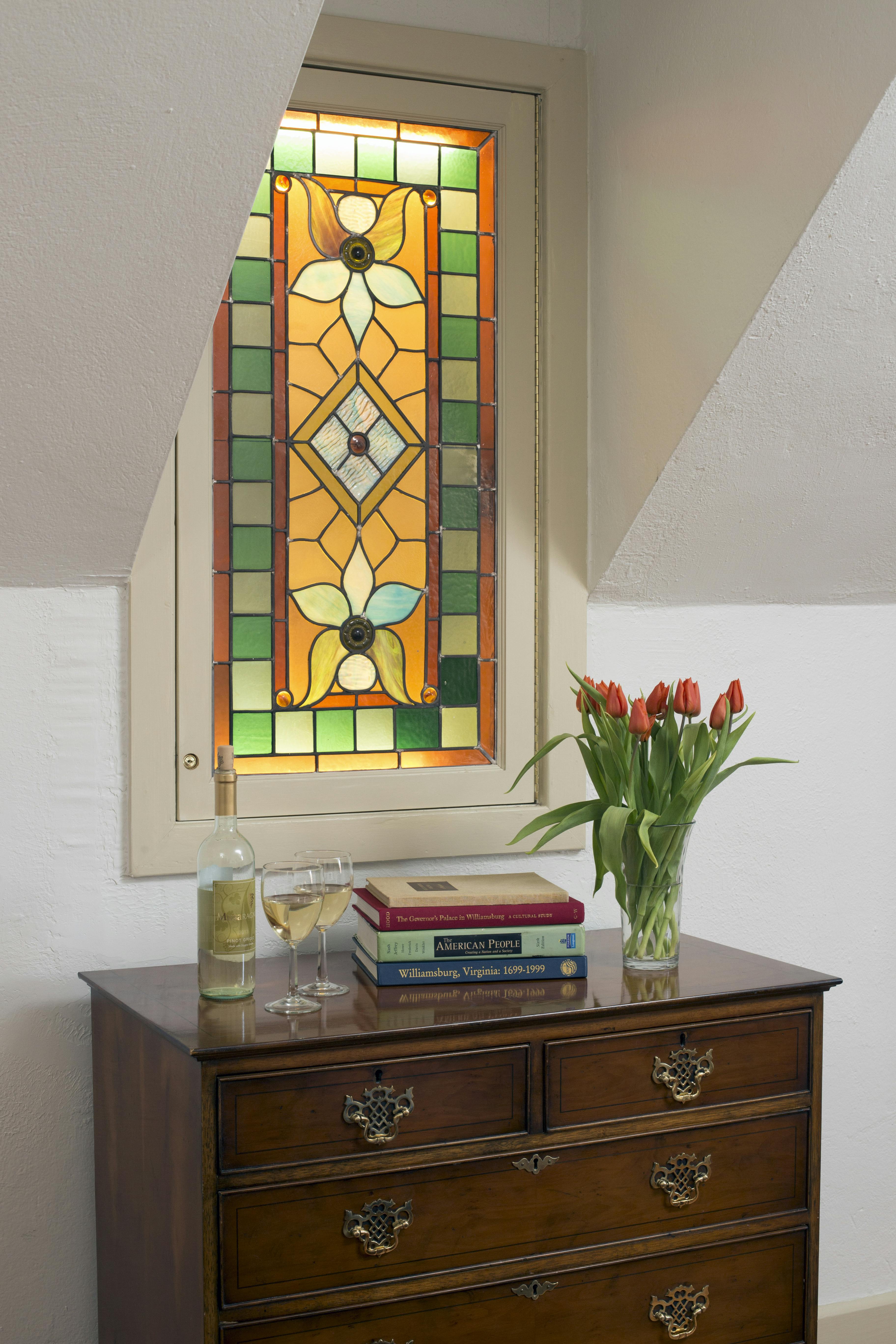 Antique chest of drawers with books, two wine glasses, a glass vase filled with tulips, and a stained glass window with flowers and geometric shapes above