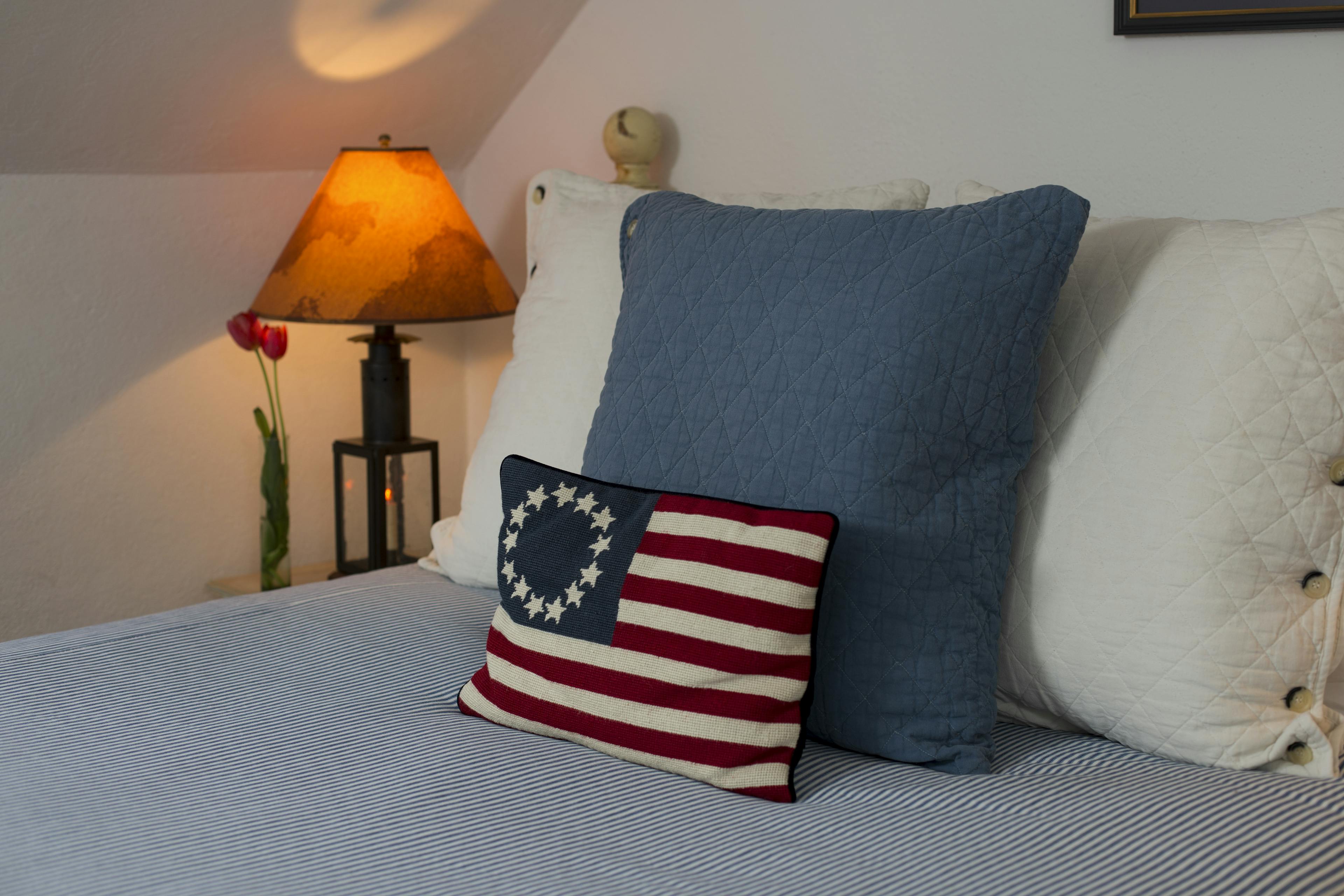 Close up of head of bed with blue & white throw pillows and a decorative colonial flag pillow, nightstand with lamp
