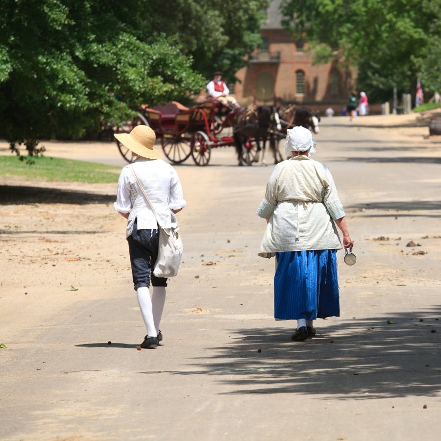 A man and woman dressed in 18th century clothing walking down a dirt street with a horse and buggy coming towards them.