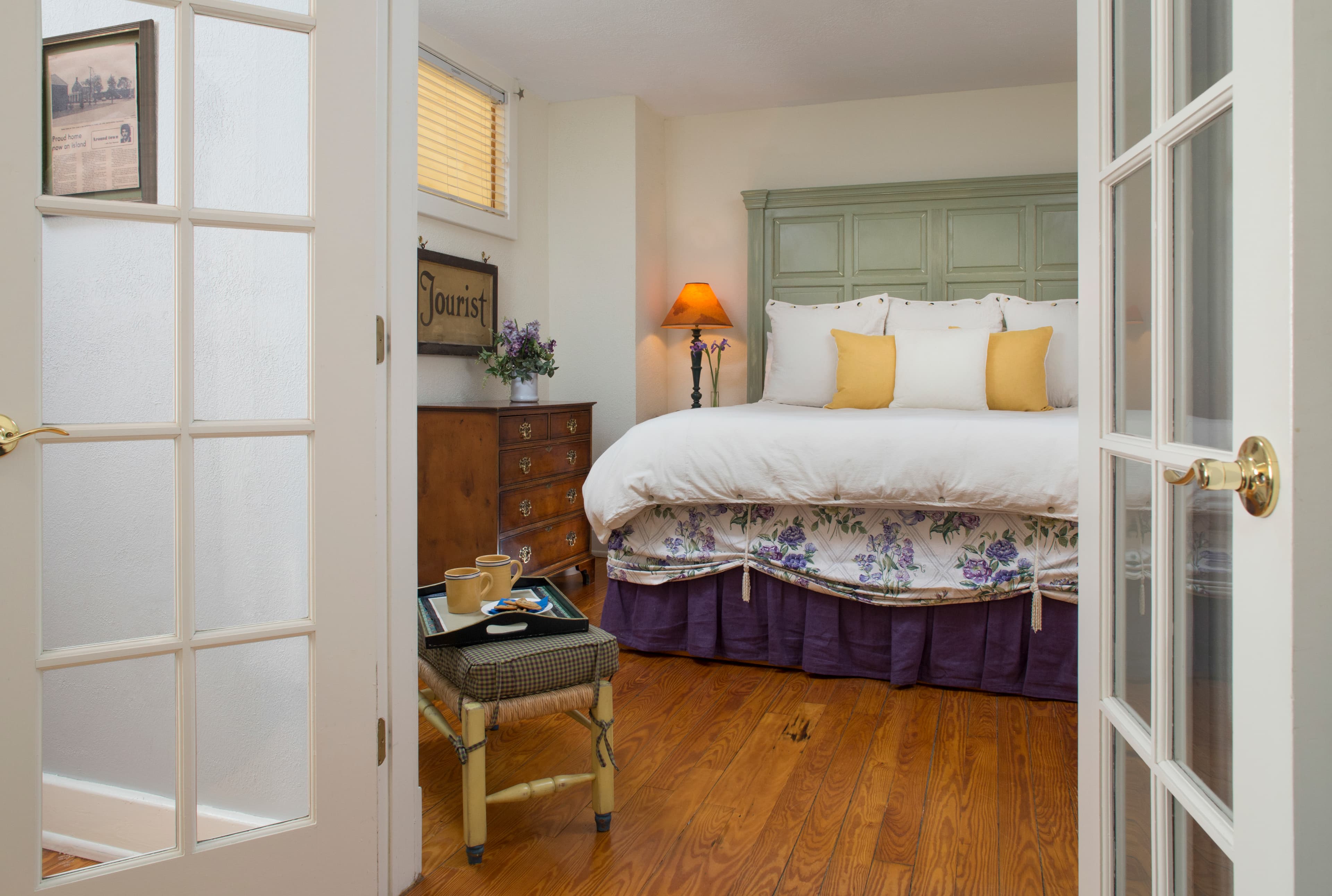 View from doorway into bedroom, beautifully appointed bed with high painted headboard, nightstand with lamp, dresser