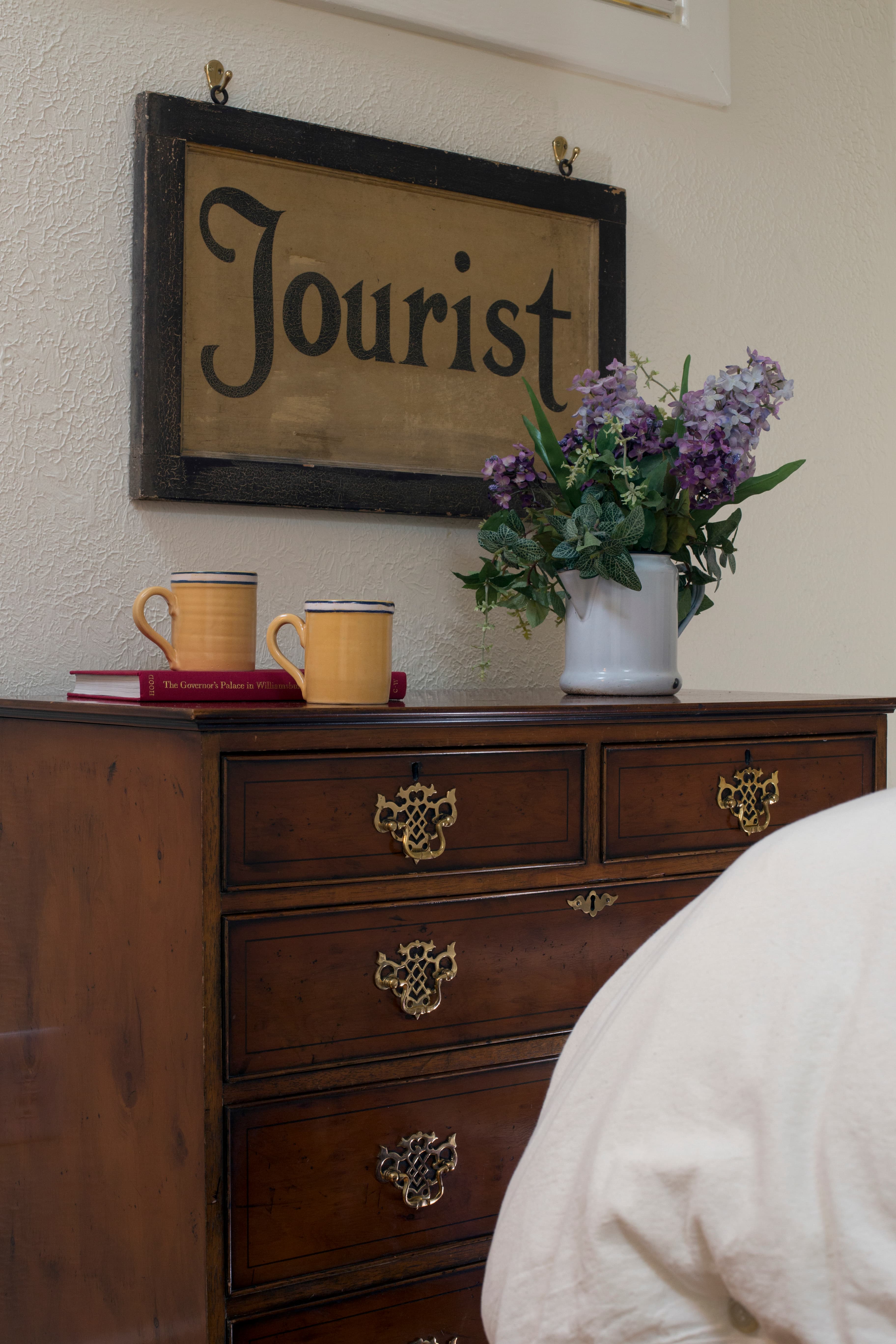 Antique chest of drawers with two mugs, a book, a vase with flowers and a framed sign reading "Tourist" hanging on wall above