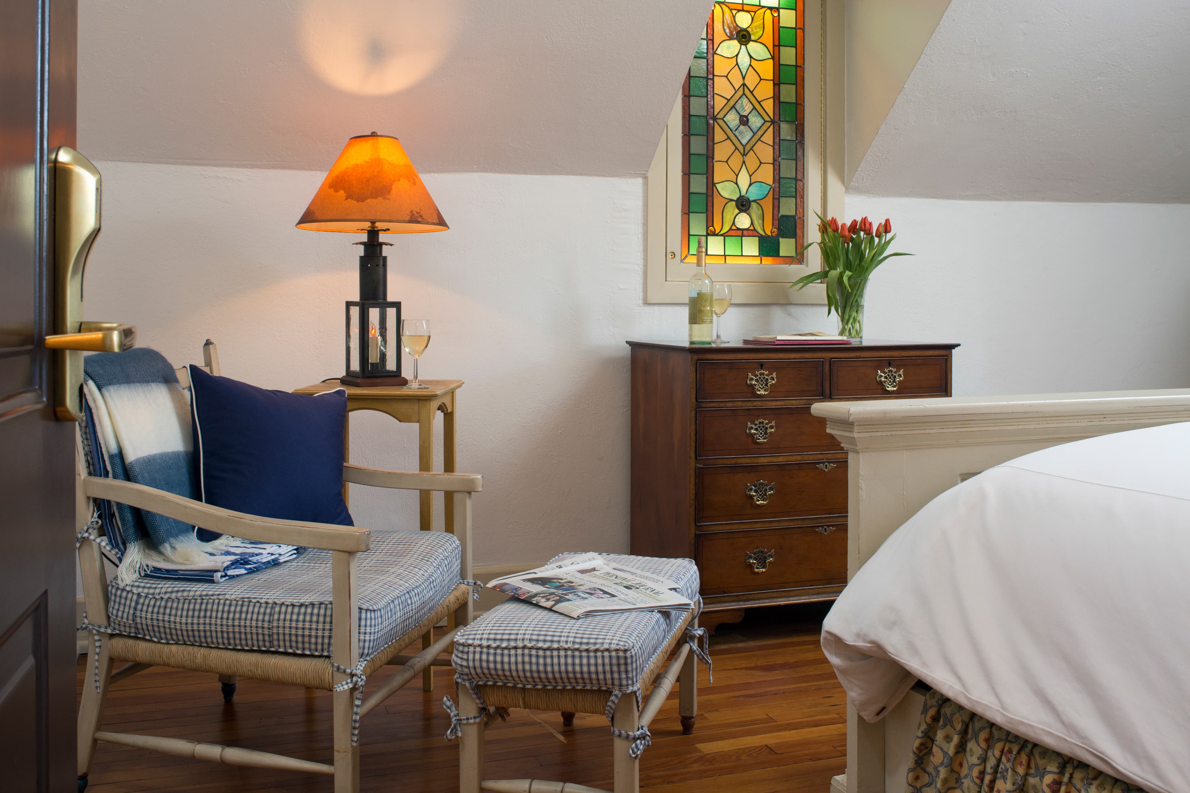 Painted wooden chair with cushions and matching footstool, side table with lamp, bureau with stained glass window above