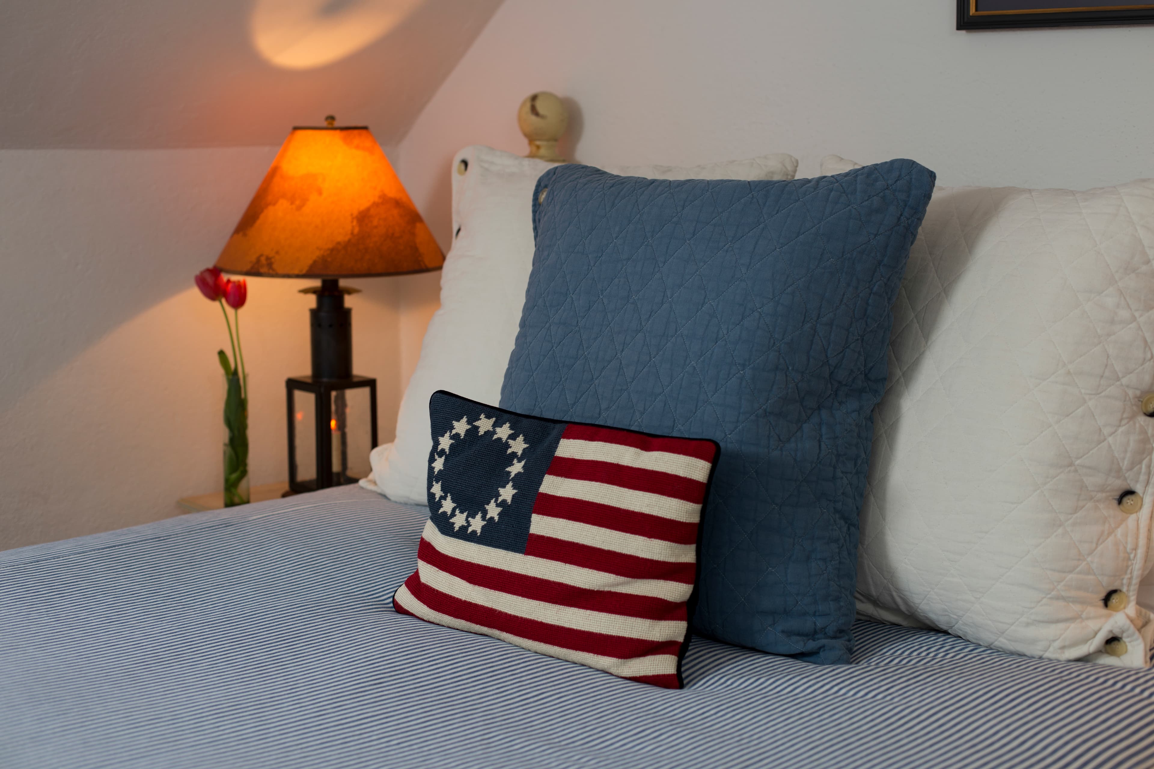 Close up of head of bed with blue & white throw pillows and a decorative colonial flag pillow, nightstand with lamp