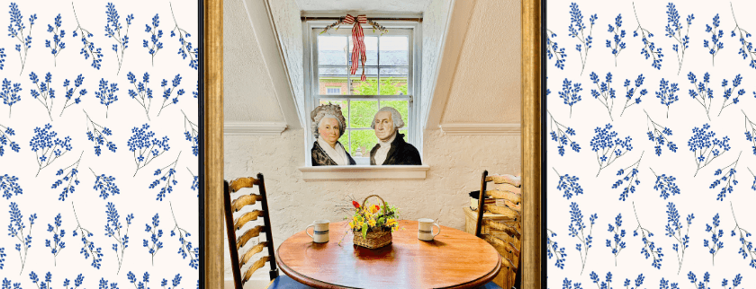 A cozy dining nook features a wooden table with a centerpiece, framed portraits of historical figures on the wall, and floral wallpaper.