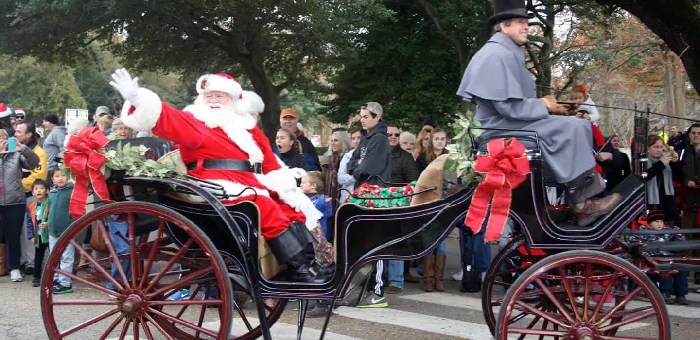 Santa Claus waves from a festive carriage during a holiday parade, surrounded by a crowd of onlookers.
