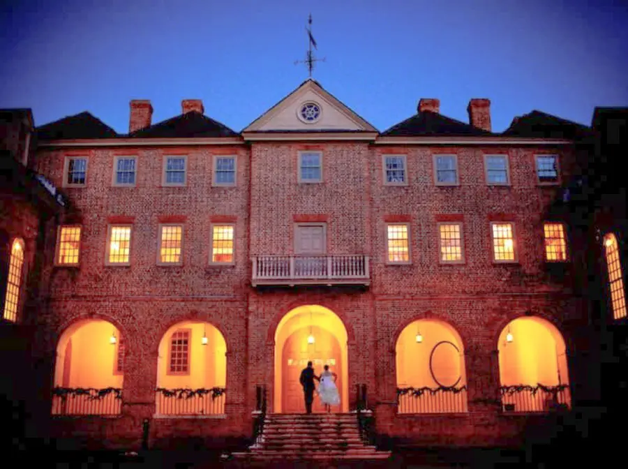 A couple stands hand-in-hand in front of a warmly lit historic building at dusk.
