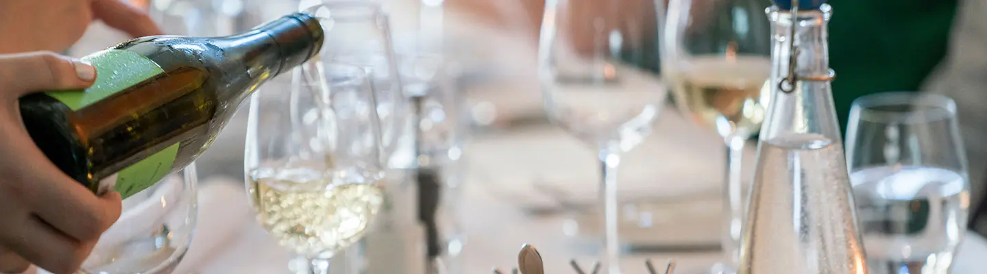 A hand pours white wine into a glass at a dining table set with several glasses.