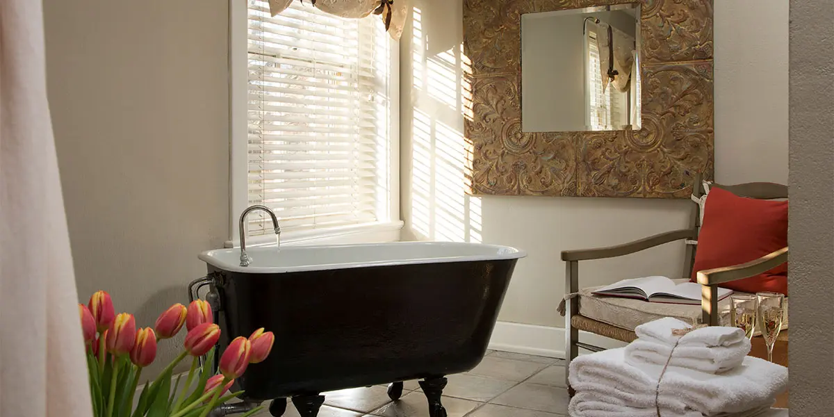 A cozy bathroom featuring a vintage black bathtub, a chair with cushions, a mirror, and fresh tulips nearby.