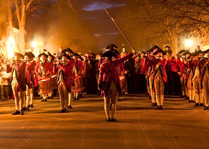 Reenactors in colonial uniforms march down a street lit by torches, accompanied by a cheering crowd. Reenactors in colonial uniforms march down a street lit by torches, accompanied by a cheering crowd.