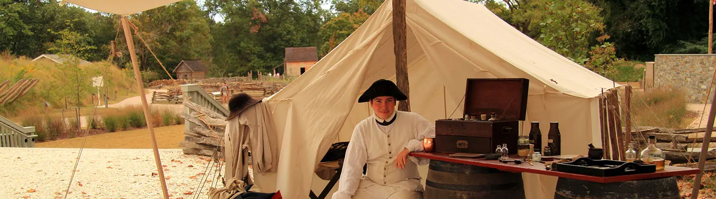 A person in historical attire stands by a makeshift table outside a tent, with a rustic camp setting in the background. A person in historical attire stands by a makeshift table outside a tent, with a rustic camp setting in the background.