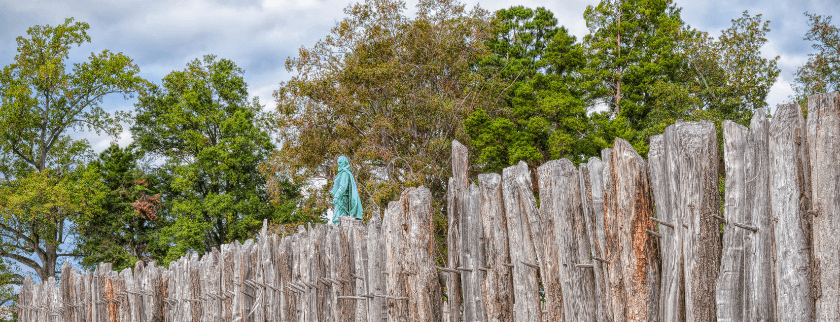A weathered wooden fence with a statue of a figure in a blue robe, surrounded by lush trees and a cloudy sky.
