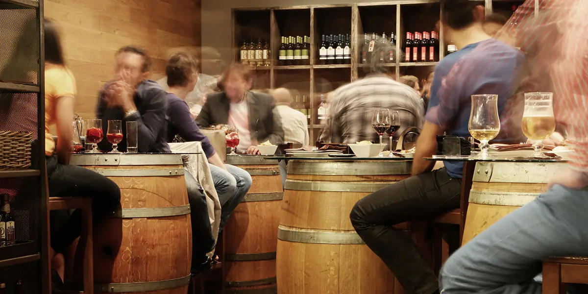 A busy bar scene with patrons seated at wooden barrel tables.