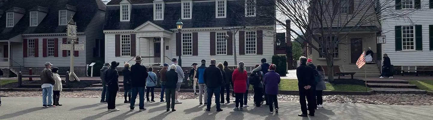 A group of people stands on a street in front of historic houses, listening to a speaker.