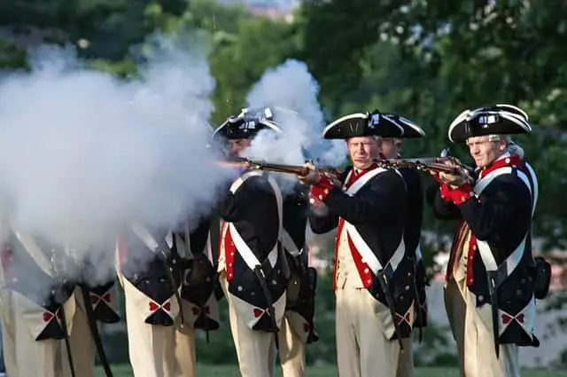 A group of historical reenactors in colonial uniform fires muskets, creating smoke.