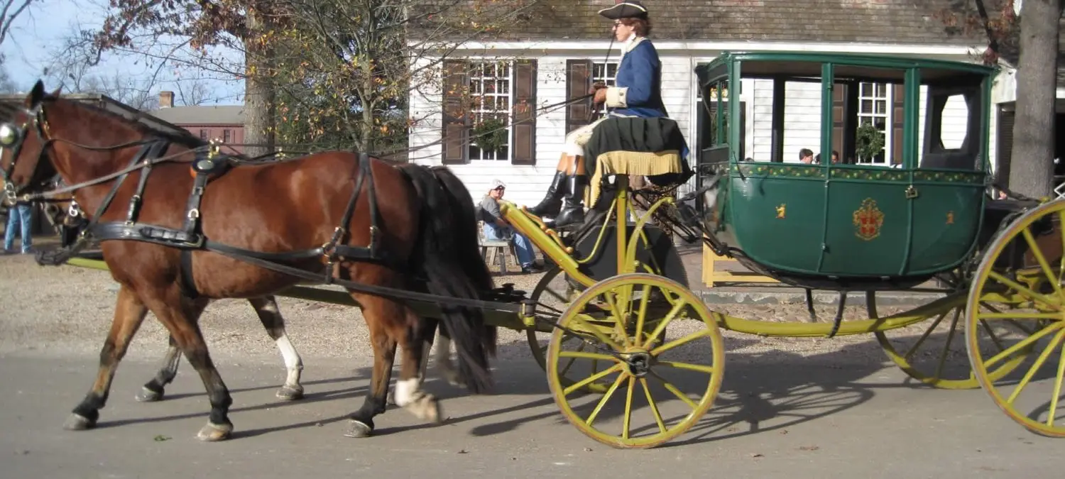 A horse-drawn carriage with a driver in historical attire moves along a road near a building. A horse-drawn carriage with a driver in historical attire moves along a road near a building.