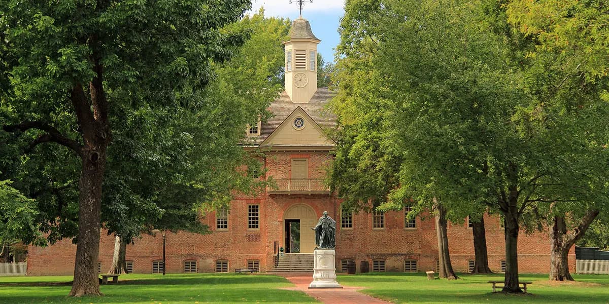 Colonial brick building surrounded by trees and a statue in a grassy area.