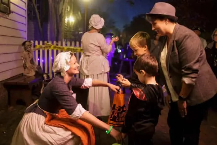 A costumed woman hands candy to two children during a Halloween event, with other costumed figures in the background. A costumed woman hands candy to two children during a Halloween event, with other costumed figures in the background.