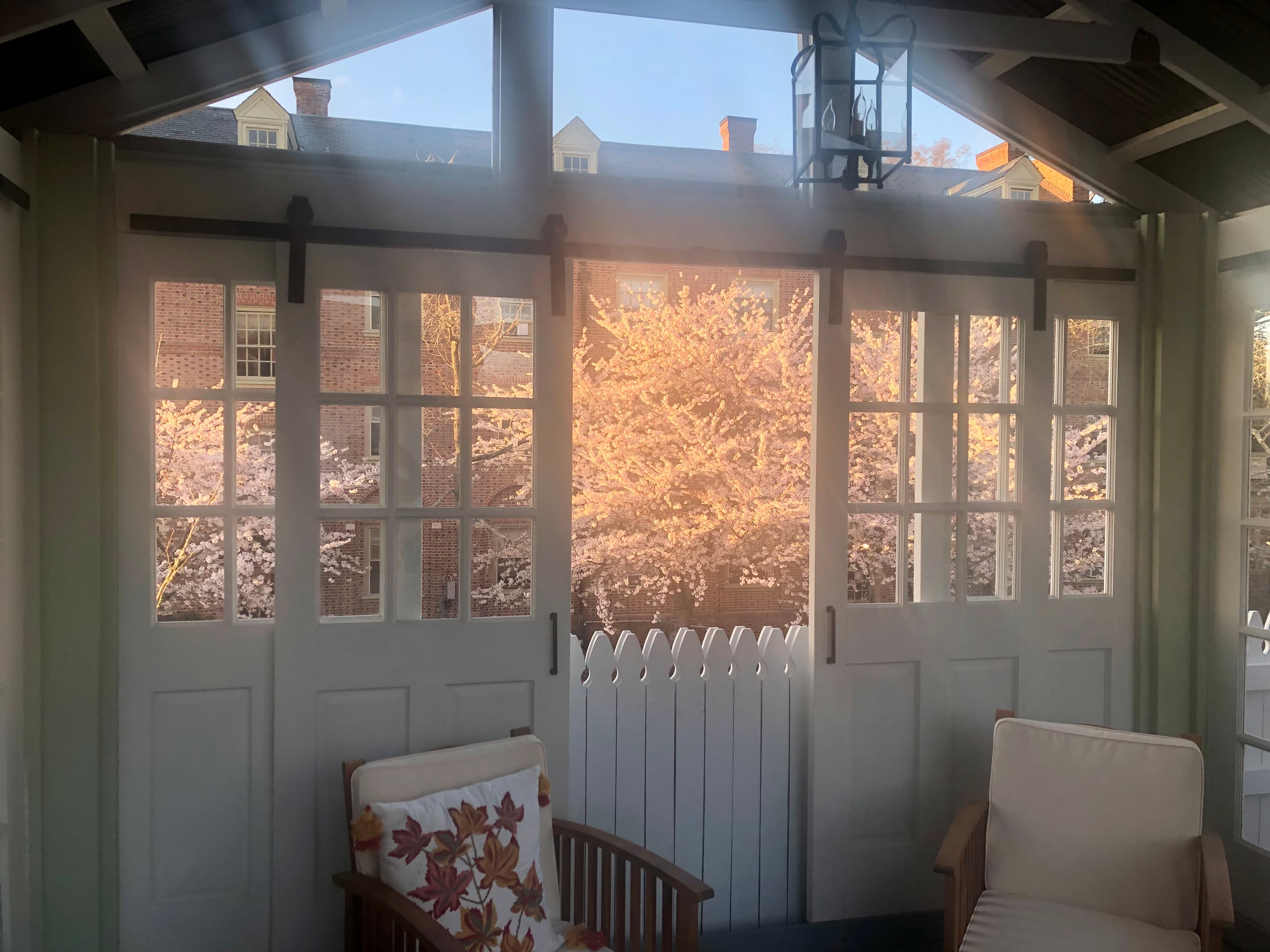 A sitting room inside white barn doors leading outside to a yard with a flowering bush and a brick building behind it.