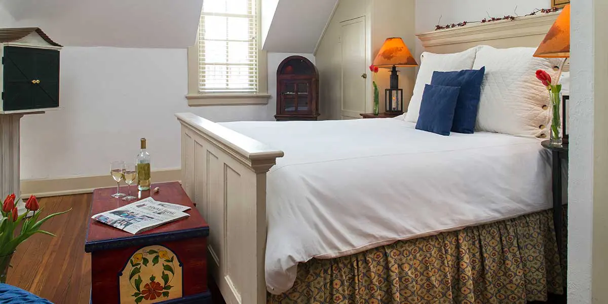A cozy bedroom featuring a neatly made bed, bedside lamps, a wine bottle with glasses, and decorative accents.