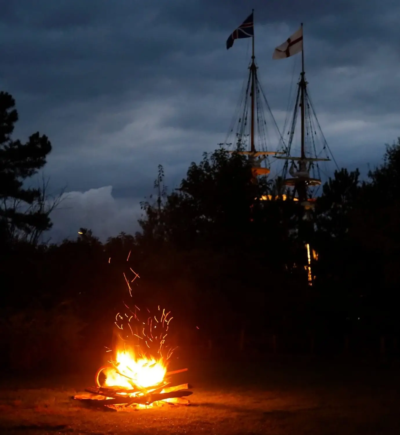 A glowing campfire illuminates a dark landscape with a ship's masts and flags in the background.