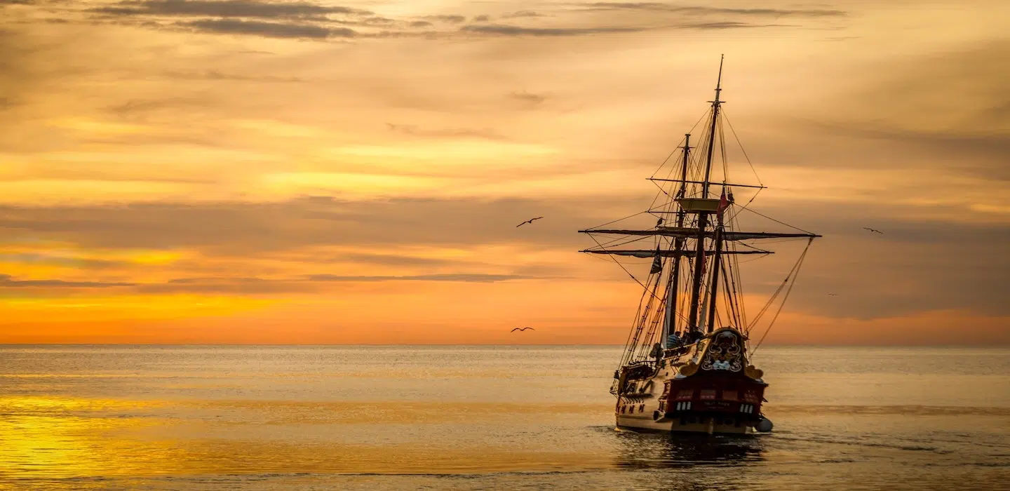 A sailing ship navigates calm waters under a vibrant sunset.