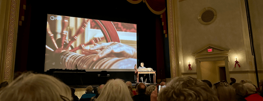A speaker presents on stage while a close-up video plays in the background, showing hands at a spinning wheel, in a theater filled with an audience.