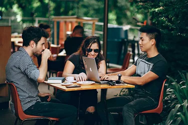 Three people collaborate at an outdoor café, working on a laptop and enjoying coffee.
