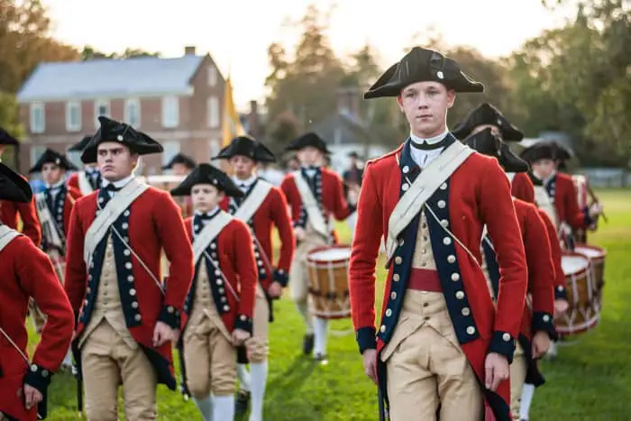A group of soldiers in 18th-century uniforms march in formation on a grassy field.