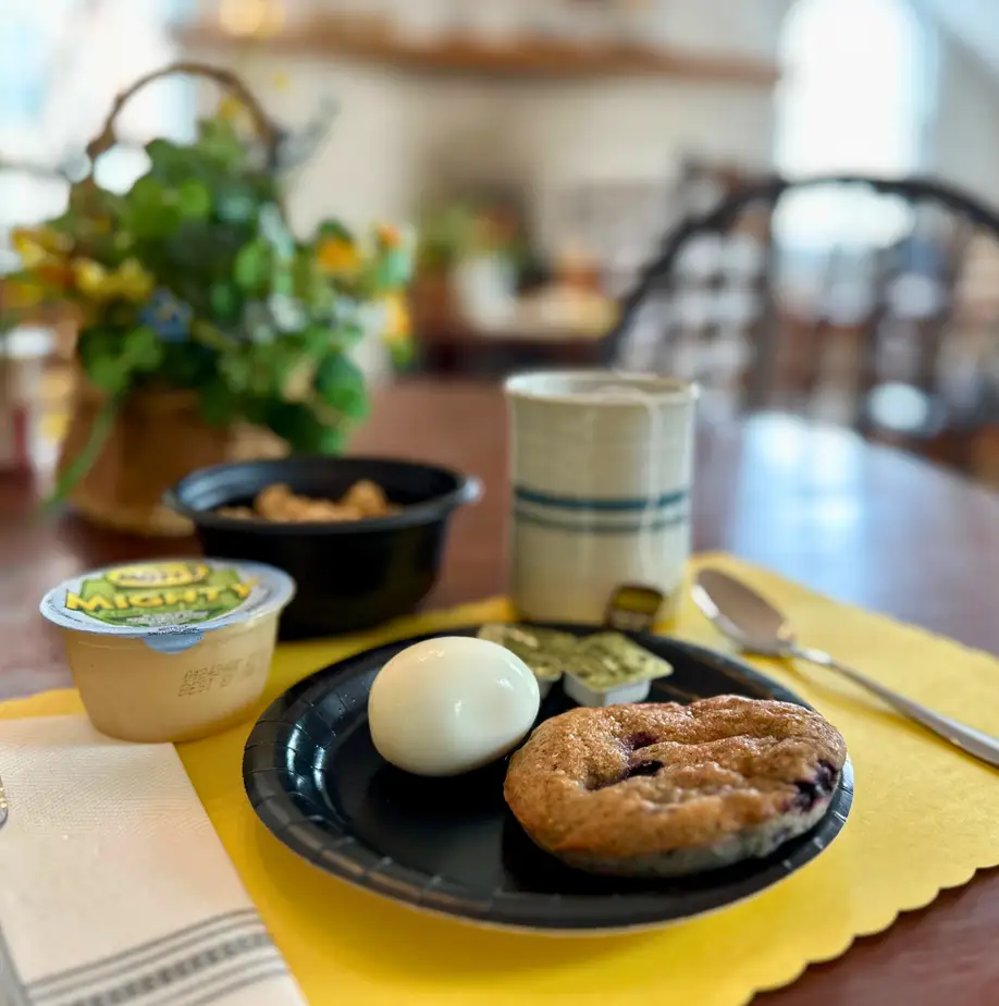 Close-up of a breakfast table setting with a boiled egg, muffin, yogurt, granola, and a cup of coffee, set on a yellow placemat.