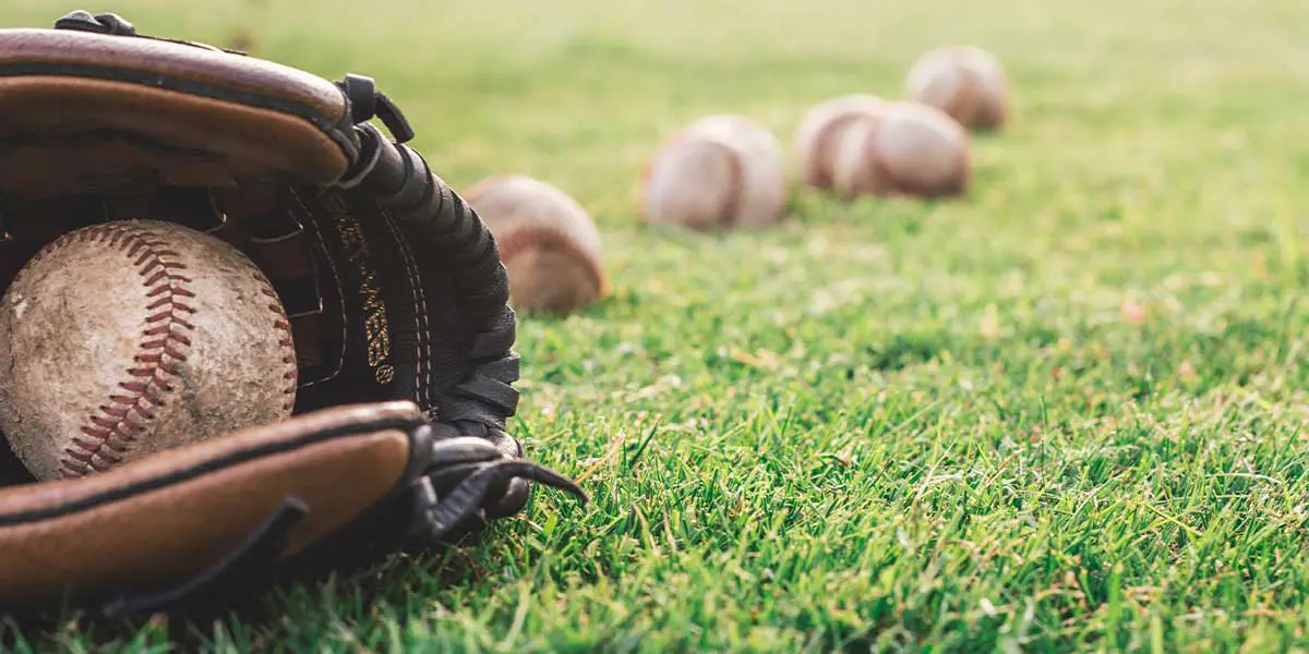 A baseball rests in a glove on grass with several more baseballs scattered nearby.