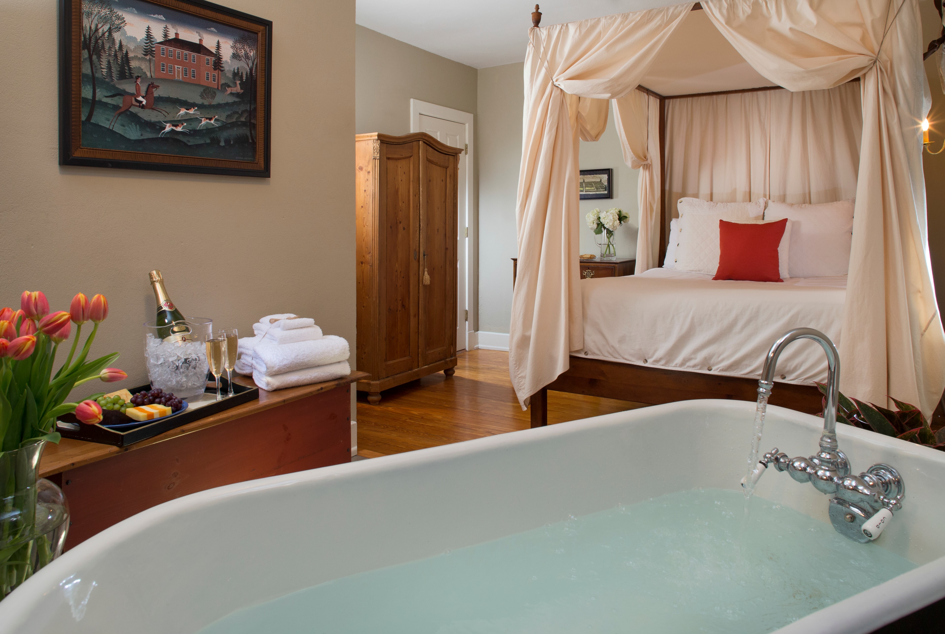 Soaking tub in foreground with vase of tulips, tray with champagne and towels nearby, in the background a luxuriously canopied four-poster bed and wooden armoire