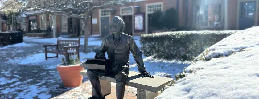 A bronze statue of a seated boy writing on a bench, surrounded by a snowy landscape and brick buildings.