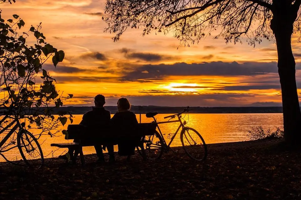 A couple sits on a bench by the water, watching a colorful sunset with bicycles nearby.