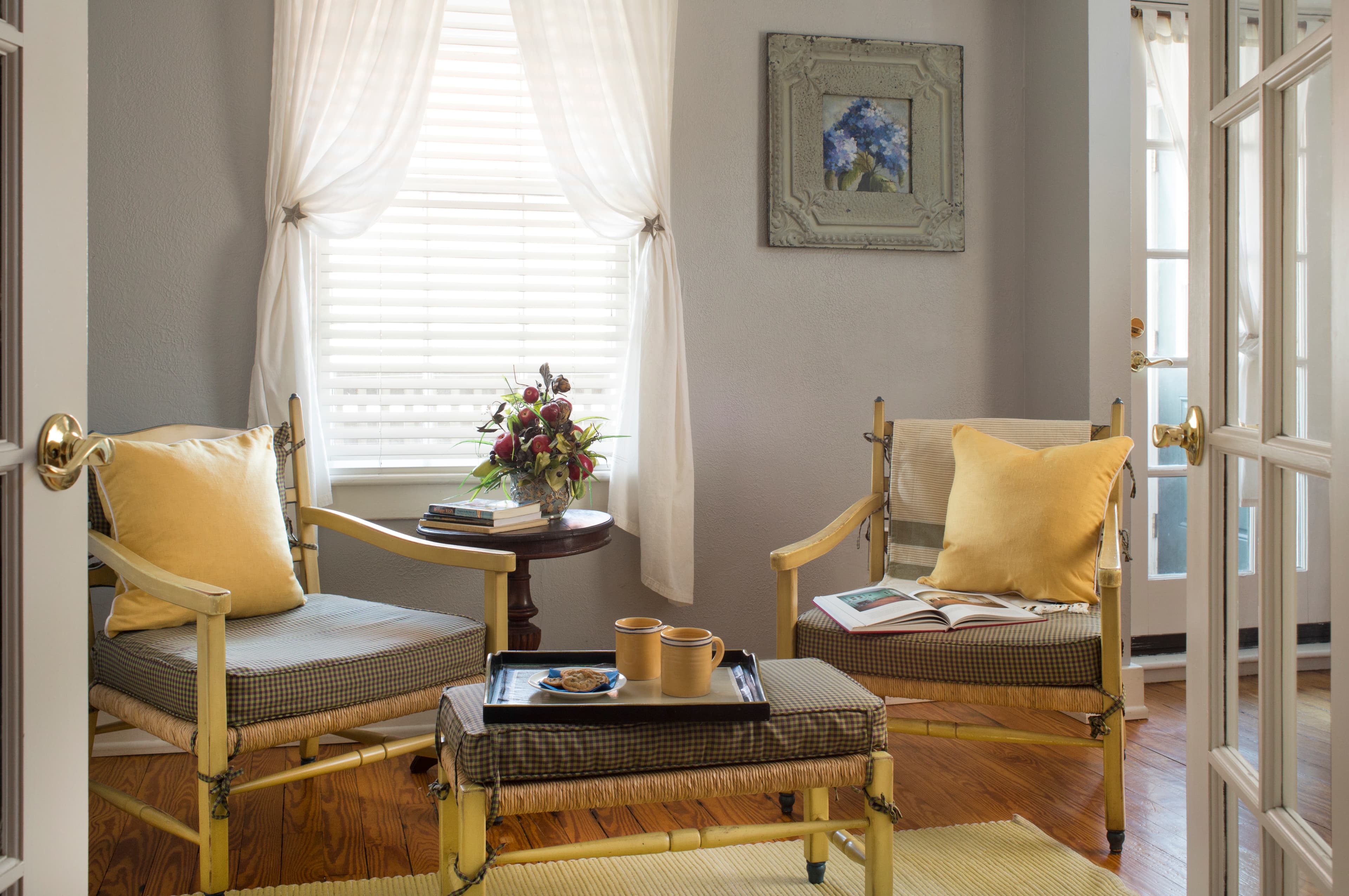 A sitting area with wood chairs with comfy cushions, a small table in between with a tray of cookies and coffee mugs, and a table in front of a window with a stack of books and flowers.