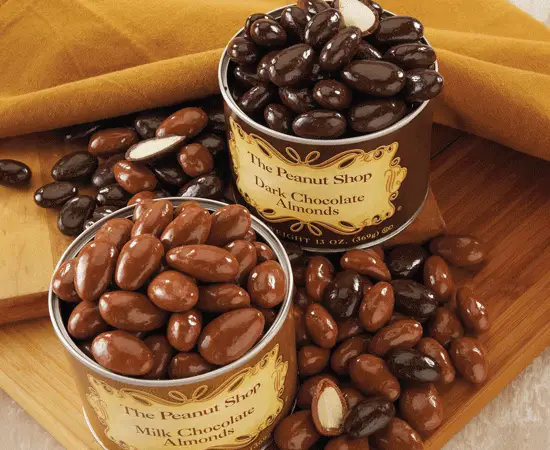 A display of two cans filled with milk and dark chocolate-covered almonds, surrounded by scattered nuts.