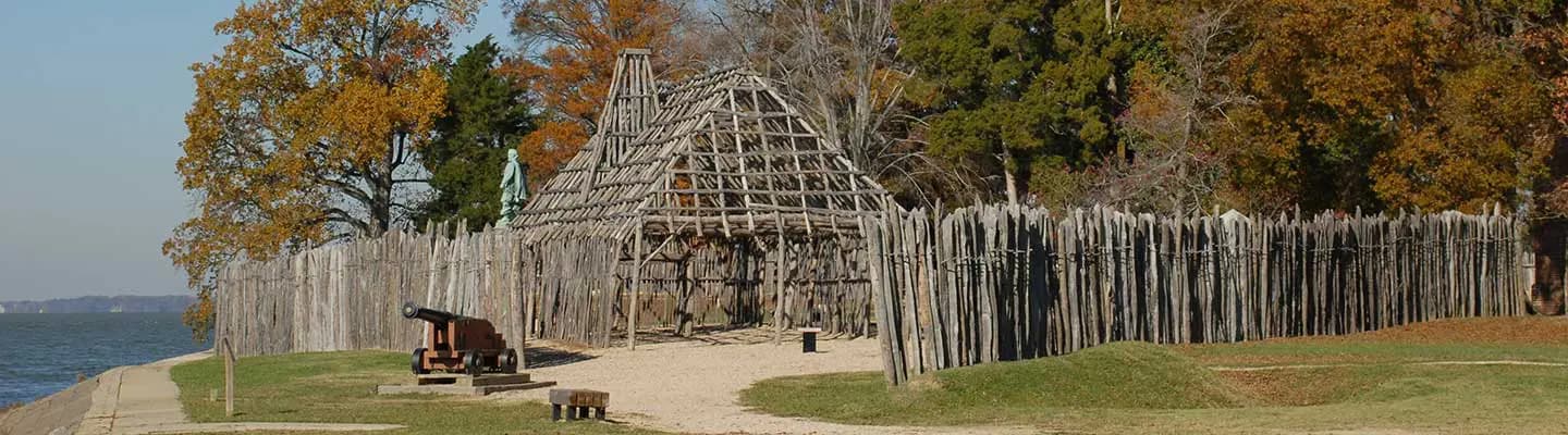 Reconstructed historic structure with a wooden palisade near a body of water, surrounded by autumn foliage. Reconstructed historic structure with a wooden palisade near a body of water, surrounded by autumn foliage.