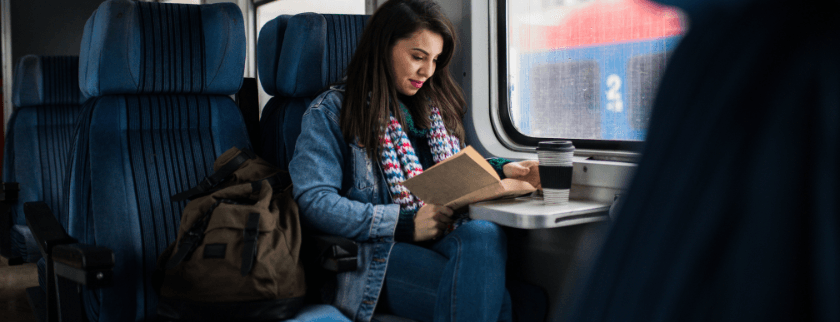 A woman in a denim jacket reads a book while seated in a train, with a coffee cup beside her.