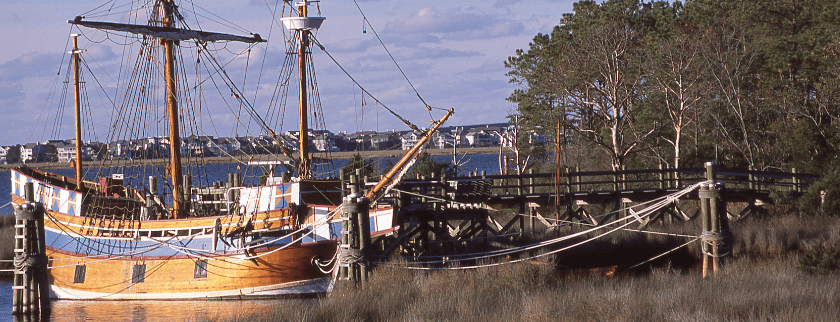 A wooden ship docked near a wooden pier, surrounded by trees and water under a blue sky.